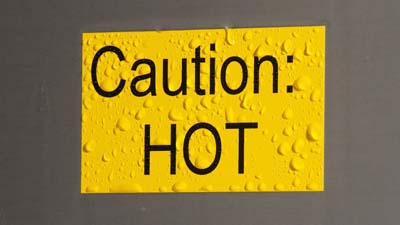 A yellow Brady toughwash label with the text CAUTION HOT printed on it. The label is wet as if it was recently sprayed clean.