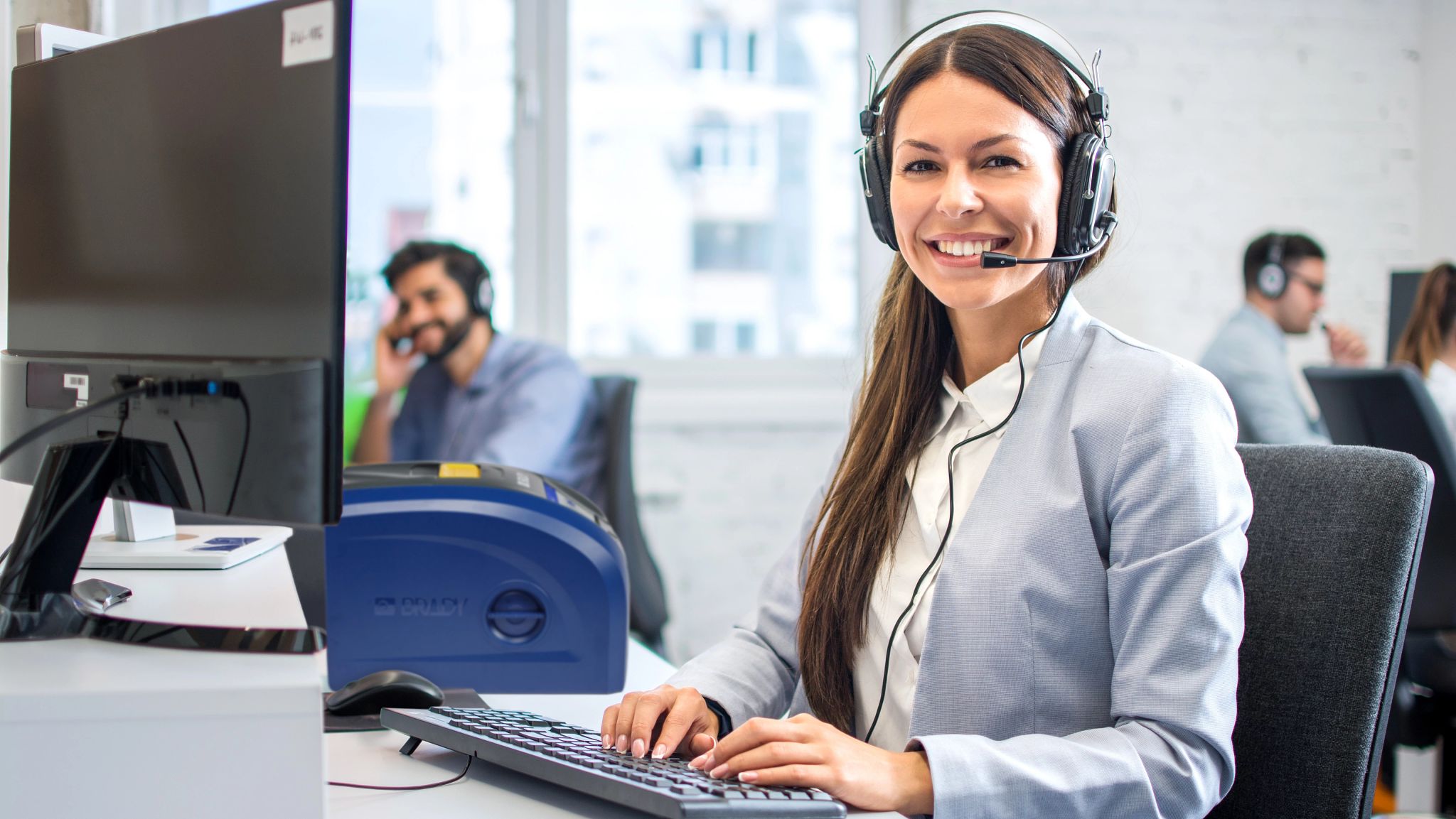 A person holding a phone. The phone screen shows a bright yellow label created using Express Labels Mobile. In the background, there is a plastic tub with the actual printed label on it.